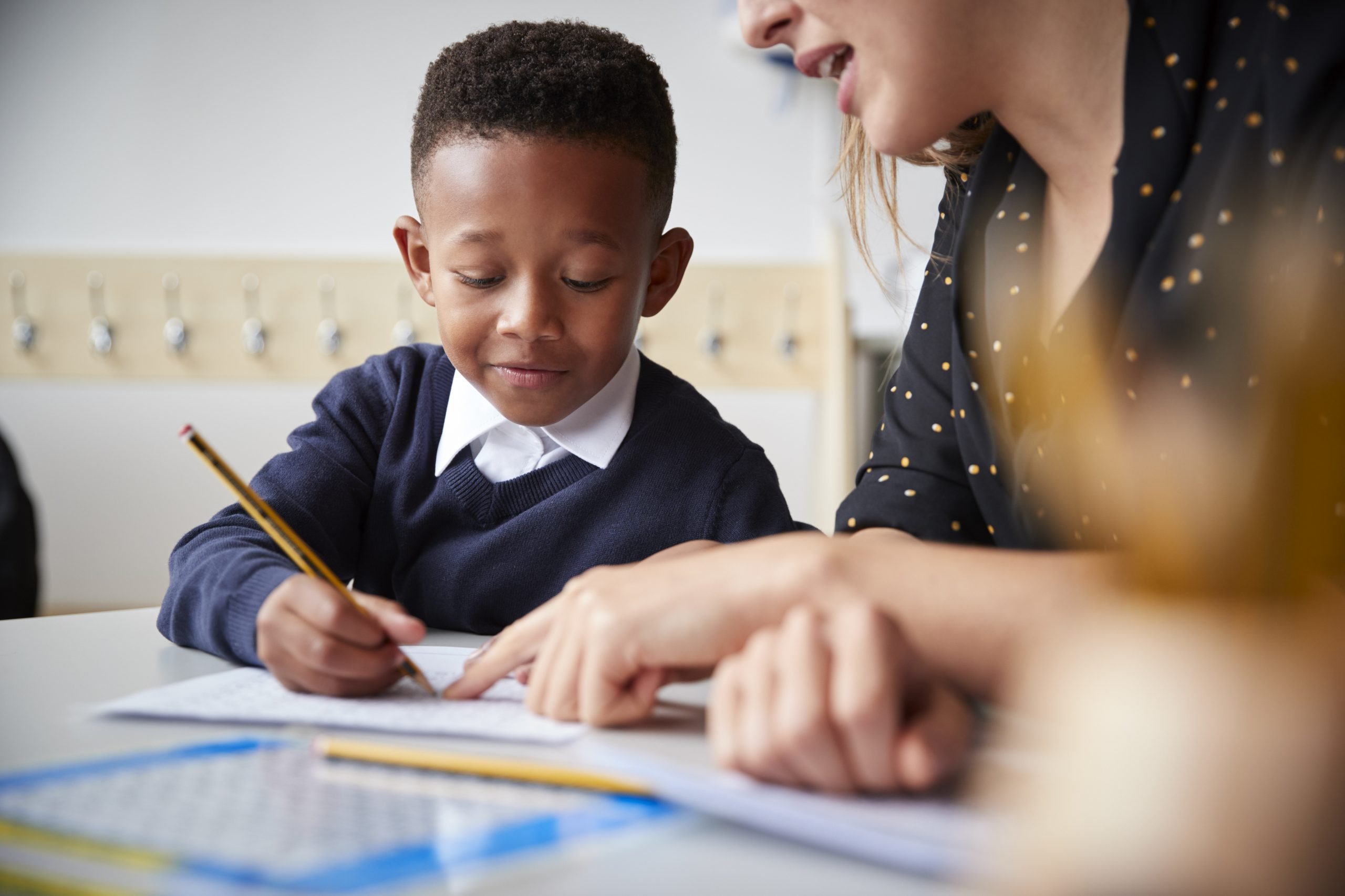 Boy in school uniform writing with a pencil under guidance of teacher.