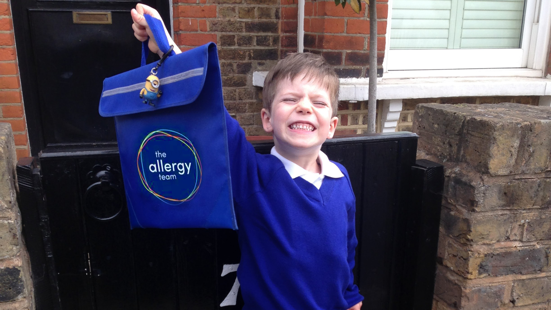 Boy in blue uniform holding up book bag and grinning